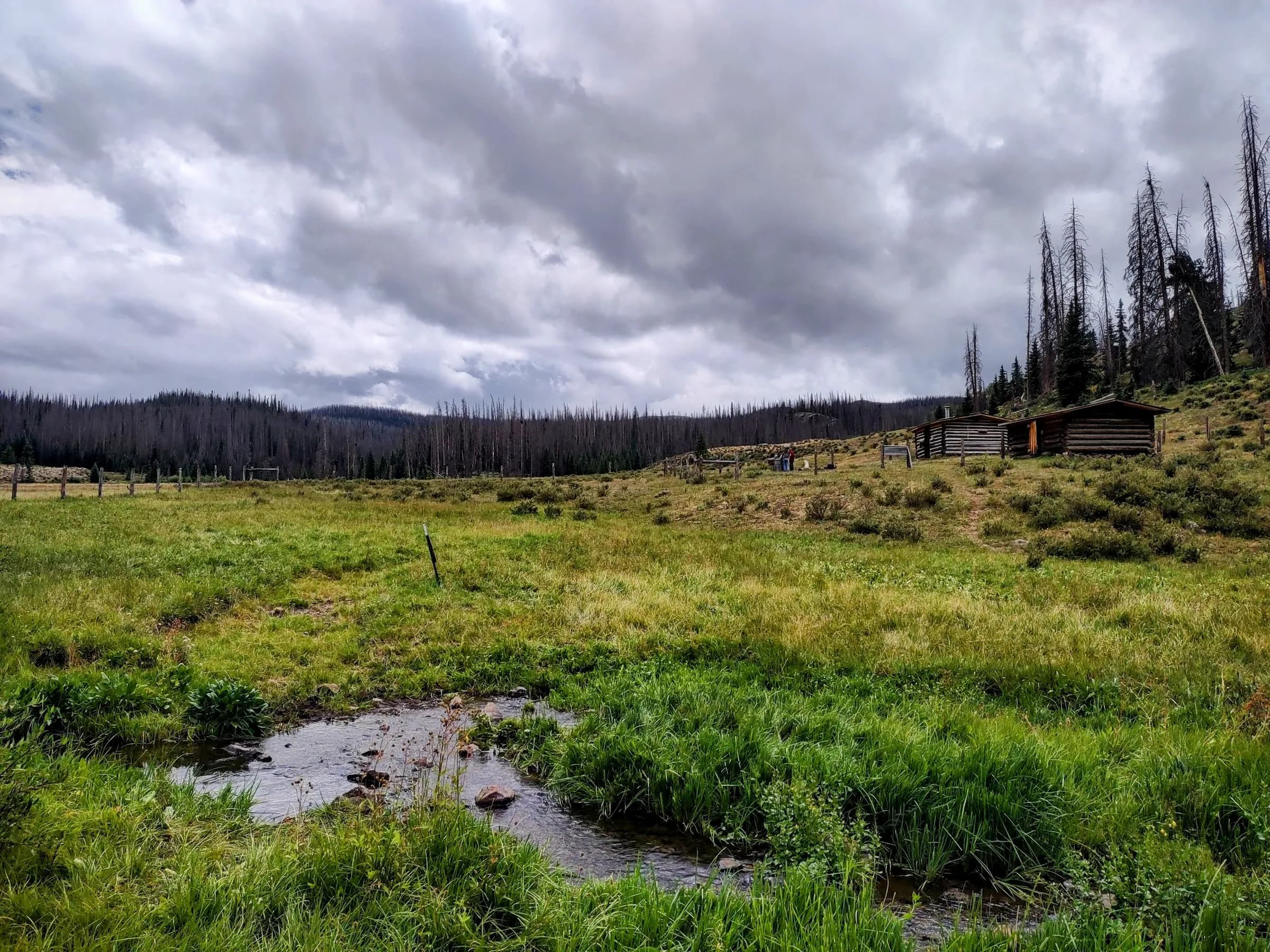 An alpine meadow with a creek and two log cabins under a stormy sky, ringed by spruce forest.